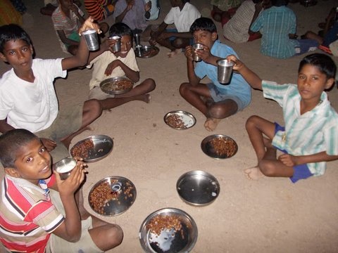 Children sitting  on dirt floor eating out of tin plates and cups. 