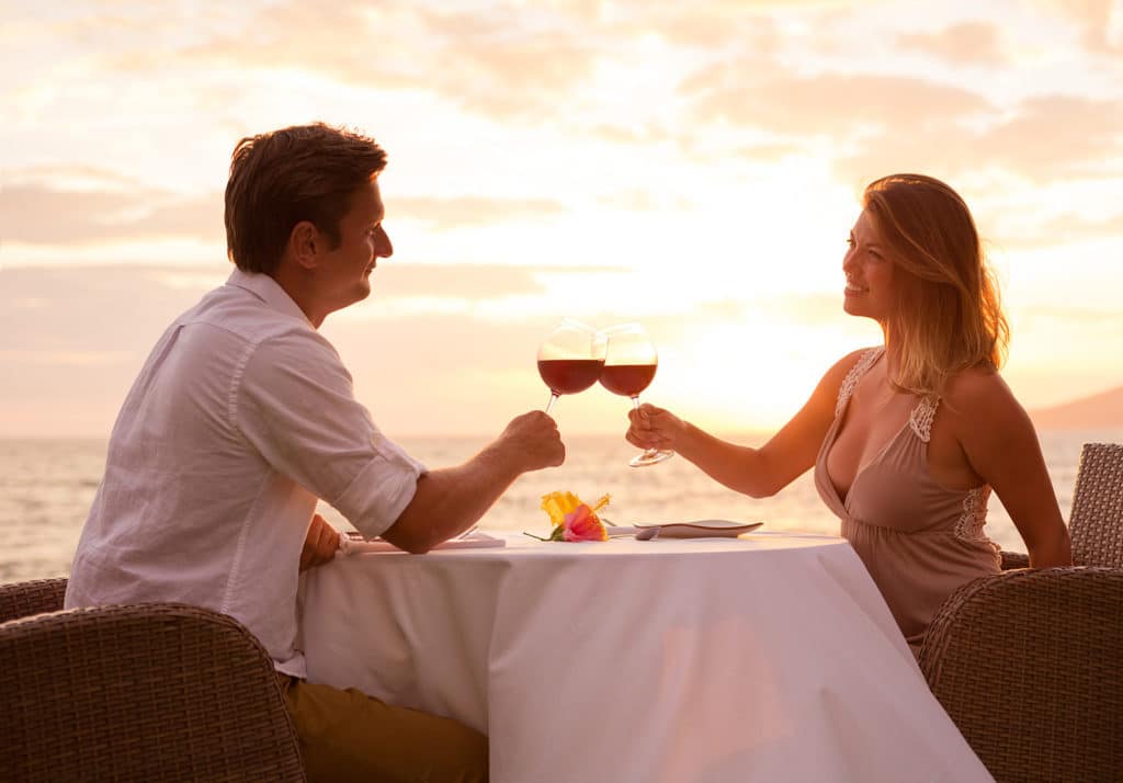 A couple toasting with wine glasses at a restaurant table overlooking an ocean sunset.